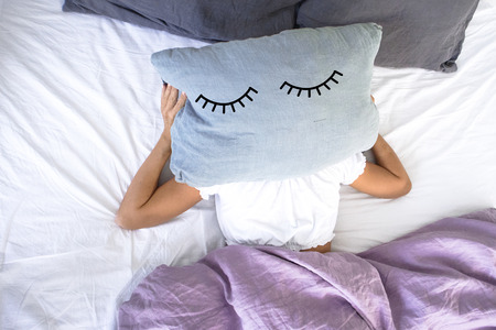 a young girl sleeping in white bed doesn't want to get up early in the morning,covering her face with a pillow with closed eyes on it.の写真素材