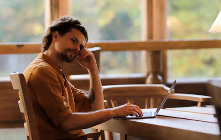 Young handsome man sitting thoughtfully at the table working and typing on laptop.の写真素材