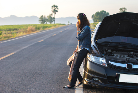 Young woman using mobile phone while looking at broken down car on streetの写真素材