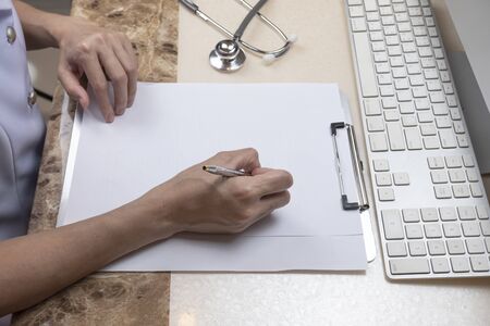 High angle view of Asian female doctor or nurse is writing something on clipboard with stethoscope and part of computer keyboard on the table at hospital office roomの写真素材