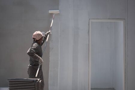 Rear side view of Asian builder worker using long handle roller brush to applying primer white paint on cement wall background inside of house construction siteの写真素材