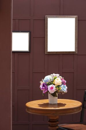 Artificial rose flowers in ceramic vase on wooden round table with teak wood chair and the old blank picture frame on wooden wall with blur door frame in foreground, vintage style and vertical frameの写真素材
