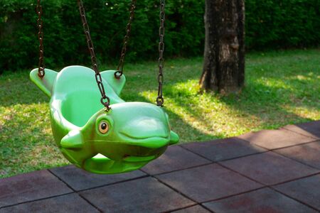 The old empty green hanging swing in dolphin shape and rubber floor tiles with green lawn in outdoor playground area at public parkの写真素材