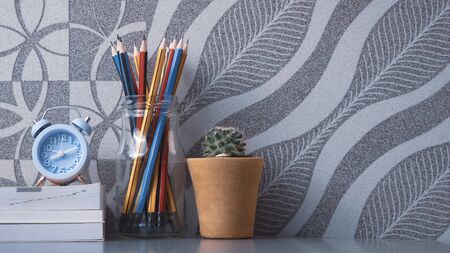 Many wooden pencils in glass jar, little alarm clock on stacked books with green cactus in clay pot...