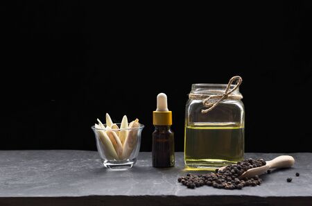 Dropper bottle with herbal essential oil in glass jar and lemongrass slice in little glass with black peppercorns in wooden spoon on black slate plate in dark backgroundの写真素材