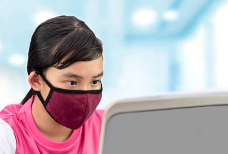 Young Asian girl in pink t shirt with red cotton face mask using laptop computer to online learning with blurred blue interior room background, new normal life after covid-19 pandemicの写真素材