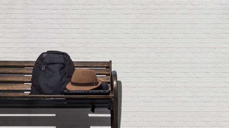 Vintage fedora hat with laptop and black backpack on metal park bench in front of white brick wall background in public park areaの写真素材