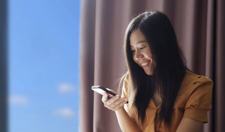 Soft image of happy beautiful Asian woman smiling and reading messages on her smartphone in living...