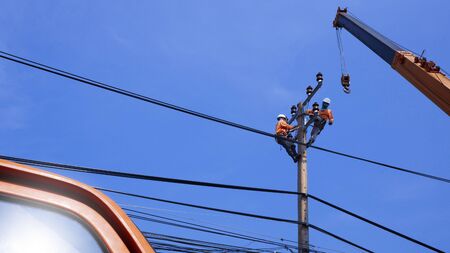 Low angle view of 2 electricians with crane truck are working to install electrical system on electric power pole against blue sky backgroundの写真素材