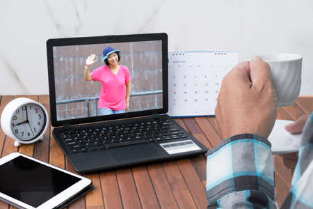 Close up man drinking hot coffee and having video call with his Asian girlfriend on laptop computer...