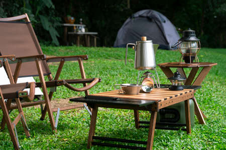 Stainless steel kettle, portable gas stove, bowl and vintage lanterns with outdoors table set on green lawn in camping areaの写真素材