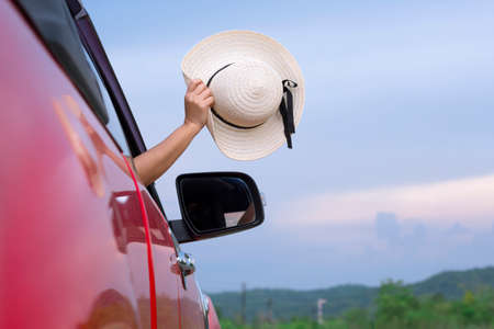 Asian female hand sticking out of red car window and waving her wide brim hat with blurred mountain and blue sky backgroundの写真素材