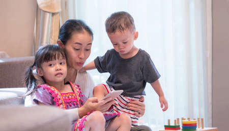 Asian mother with 2 children are relaxing on the sofa and looking smart phone with a surprised...