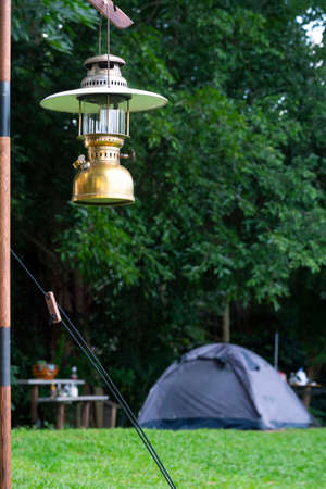 Focus at vintage camping brass lantern with blurred background of tents on green lawn in camping area at natural park in vertical frameの写真素材