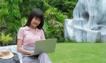 Asian female blogger using laptop to write post on social network in beautiful gardening area at...