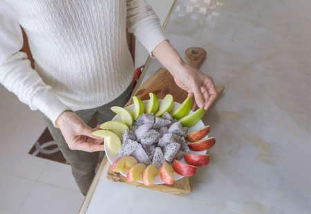 Part of woman in white long sleeved  is preparing red and green apples and dragon fruit on marble counter in the kitchen at homeの写真素材