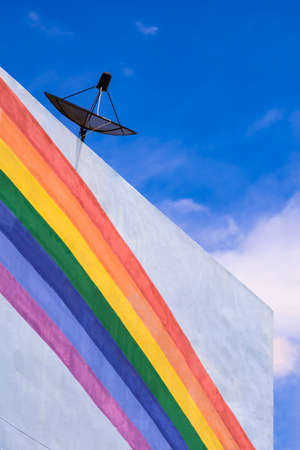 Low angle and side view of satellite dish on top of white building with rainbow stripes on building wall against blue sky backgroundの写真素材