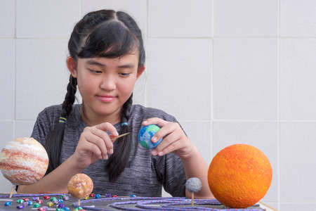Selective focus at young Asian girl making homemade solar system model on wooden table at home for her science homework project at schoolの写真素材