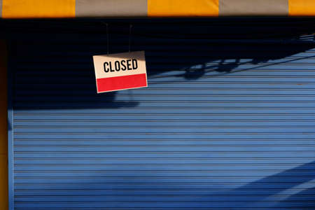 Sunlight and shadow on surface of hanging closed sign board in front of blue shutter door of store in morning timeの写真素材