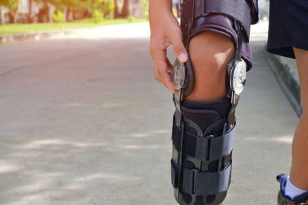 Young man's hand turns angle movement control of supportive knee brace on his leg for jogging with flare light and blurred public park backgroundの写真素材