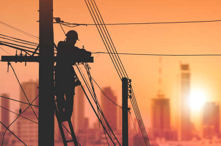 Silhouette electrician on ladder is installing cable lines to connecting internet signal on electric power pole with blurred cityscape view in sunrise sky backgroundの写真素材
