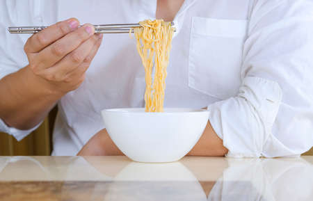 Close up man's hand in white shirt using silver chopsticks to eat instant noodles in white bowl at homeの写真素材