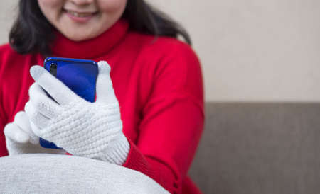 Close up image of young smiling woman in red sweater using modern smartphone to video call on sofa in living room during winter seasonの写真素材