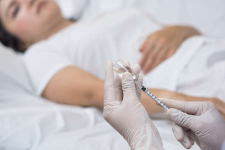 Focus at doctor's hand holding draw syringe with vial for injection into female patient on bed in hospital roomの写真素材