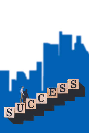 Miniature businessman walking on wooden blocks with word 'success' stacking as step stair with blurred blue cityscape shadow on white background in vertical frame, business and success idea conceptの写真素材