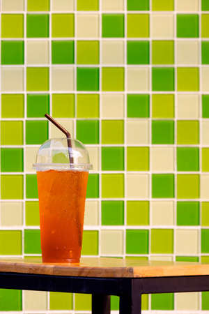 Close up of iced lemon tea in plastic glass on wooden table with blurred green glass mosaic tiles wall background in vertical frameの写真素材