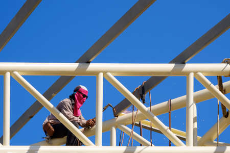 Asian construction worker is welding metal on top of building roof structure against blue sky background in sunny dayの写真素材