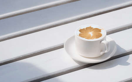 Mild sunshine on white ceramic cup of hot latte art coffee on wooden garden bench at vintage coffee shopの写真素材
