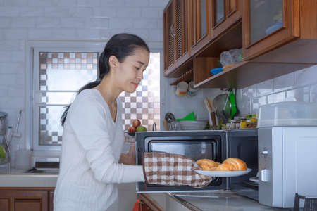 Asian woman using microwave oven to warm croissants in the kitchen at home
