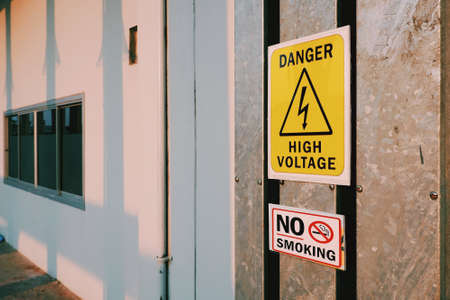 Warning danger high voltage sign and no smoking label on steel pipe cover with sunlight and electric pole's shadow on surface of white wall, sign and symbol with safety conceptの写真素材