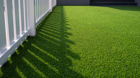 Sunlight and shadow of white wooden fence on green artificial turf surface in front yard of home, selective focus with copy spaceの写真素材