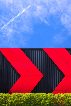 Red arrows pattern on black corrugated metal wall with green bush fence at public health park against jet plane flying on blue sky background in vertical frameの写真素材