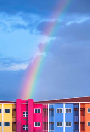 Front view of colorful apartment building against rainbow forming on blue sky after raining at...