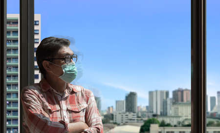 Asian man wearing protective mask and looking out of glass window at home with blurred city view...