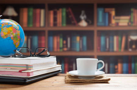 White coffee cup, eyeglasses with books stacked and black laptop on wooden table with blurred bookshelf in home office room areaの写真素材