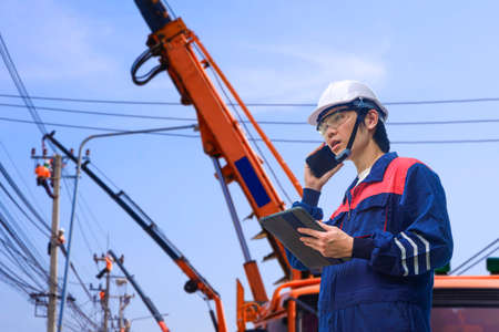 Asian engineer holding tablet and talking on smartphone while controlling his electric teamwork are working to install electrical systems on electric power poles in public areaの写真素材
