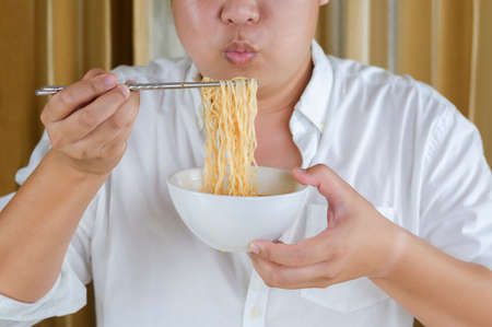 Close up of Asian plus size man in white shirt using silver chopstick to eating instant noodles at home with blowing on hot noodlesの写真素材