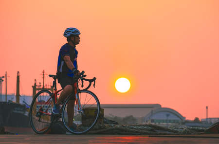 Side view of adult athletic man on bike is taking break and relaxing at harbor after long distance workout in sunset timeの写真素材