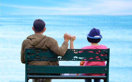 Rear view of Asian adult couple on metal bench showing pinky swear hand sign to be a relationship forever after dating on the beach, travel and love conceptの写真素材
