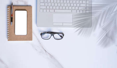 Flat lay of laptop, smartphone mockup, eyeglasses, pen spiral notebook on white marble tabletop with copy spaceの写真素材