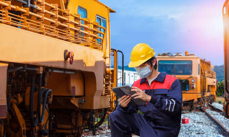 Young Asian engineer in safety workwear with protective mask using digital tablet to record information of the old trains during maintenance at locomotive maintenance station in countrysideの写真素材