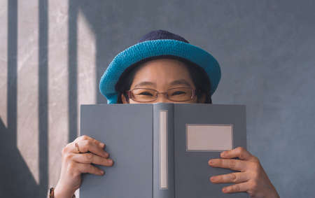 Emotional woman with eyeglass smiling and looking at camera while hiding her face behind a book against gray vintage wall backgroundの写真素材