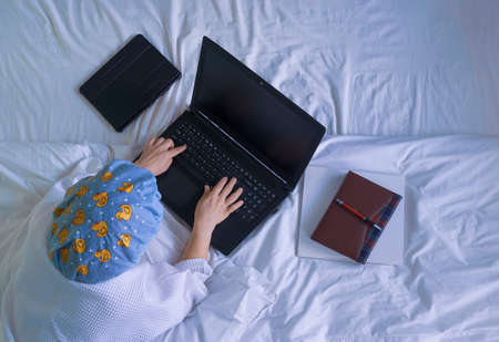 Top view of woman in white bathrobe with shower cap lying down and using laptop computer on her bed at morning timeの写真素材