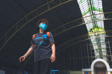 Low angle view of Asian female traveler in protective mask holding suitcase while walking and looking around at railway stationの写真素材