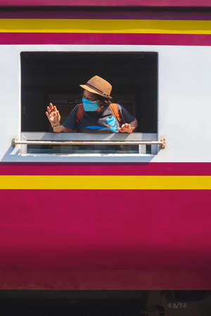 Asian female traveler wearing protective mask in train window looking sideway and waving hand while the train leaving the stationの写真素材