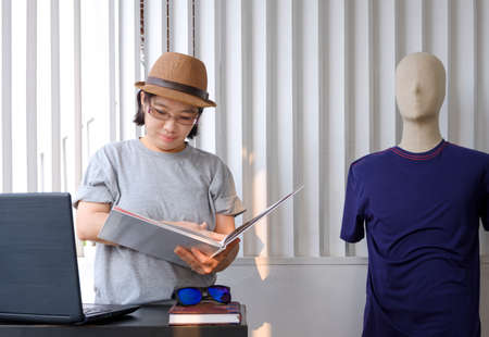 Asian plus size LGBT female designer in gray t-shirt with fedora hat is looking fashion magazine with laptop computer on the table in home office roomの写真素材